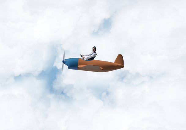 プロペラ飛行機を運転ゴーグルで飛行士の帽子をかぶった若者。飛行機のコンセプトで世界中を旅する。雲と空に小さな飛行機で飛ぶ面白い男。極端な航空趣味. - 写真・画像