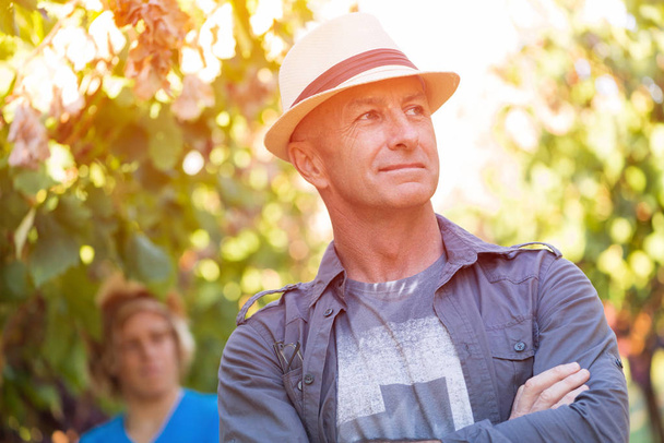 Winegrower standing in vineyard. Handsome senior man in straw hat and shirt posing. Traditional family winemaker business. Harvesting in winery industry. Man with arms folded outdoor at sunny day. - Photo, Image