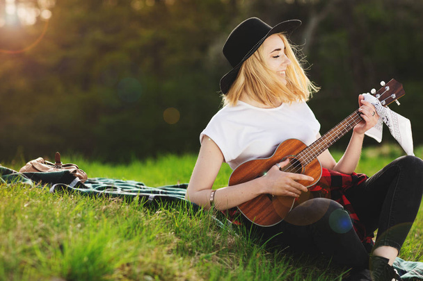 黒い帽子をかぶった若い美しい女性の肖像画。草の上に座ってギターを弾く少女 - 写真・画像