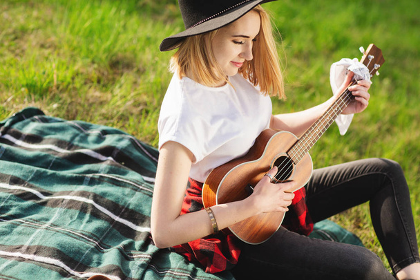 黒い帽子をかぶった若い美しい女性の肖像画。草の上に座ってギターを弾く少女 - 写真・画像