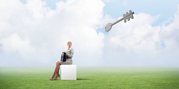Elegant businesswoman with suitcase in hand sitting on white cube and stone key symbol - Foto, imagen
