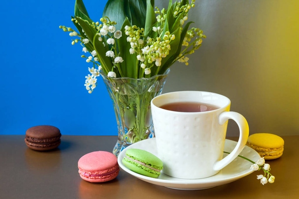 Pasteles franceses multicolores macarons y una taza de té. Un ramo de lirio del valle en un jarrón de cristal
. - Foto, Imagen