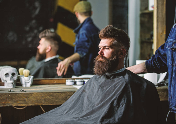 Man with beard covered with black cape sits in hairdressers chair, mirror background. Man with beard client of hipster barbershop. Barbershop concept. Hipster with beard waits for barber and haircut - Фото, зображення