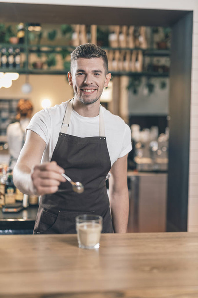 Cheerful barista at the bar counter holding teaspoon near the glass - Foto, imagen
