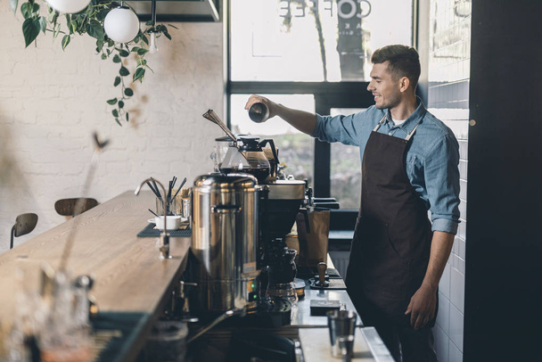 Mladý barista prosévá kávu do nádoby na obilí a usmívá se - Fotografie, Obrázek