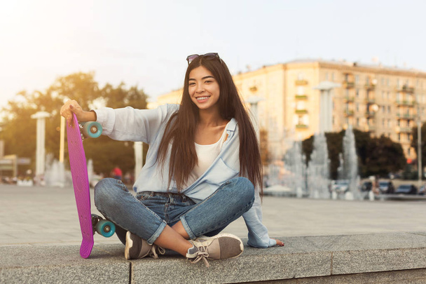 Крутая азиатская девочка-подросток, сидящая на скейтборде
 - Фото, изображение