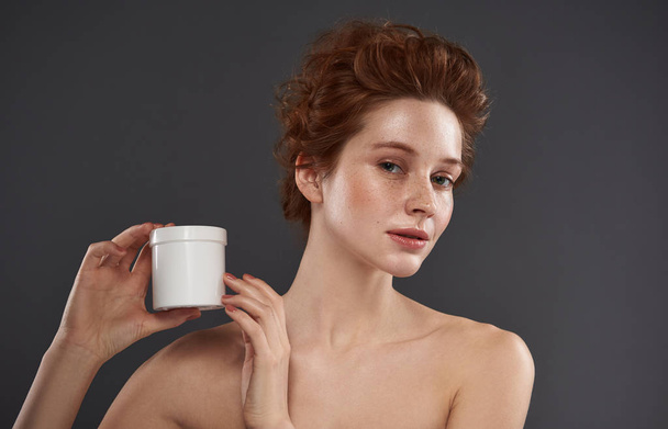 Charming red-haired girl holding white jar of cosmetic cream - Foto, afbeelding