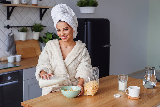 Close up jovem mulher em roupão derrama leite em prato dep com flocos de milho. Menina bonita tem um pequeno-almoço saudável na cozinha acolhedora elegante na parte da manhã
. - Foto, Imagem
