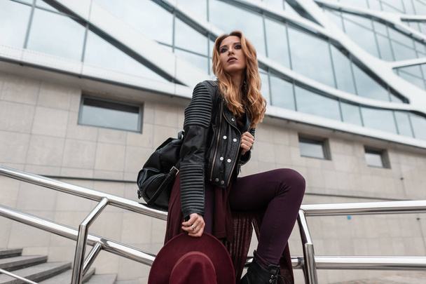 Die hübsche rothaarige junge Frau mit lila Hut in modischer Kleidung und Lederrucksack sitzt auf Metallrohren in der Nähe eines modernen Gebäudes in der Stadt. europäisches Touristenmädchen beim Ausruhen. Moderne Mode. - Foto, Bild