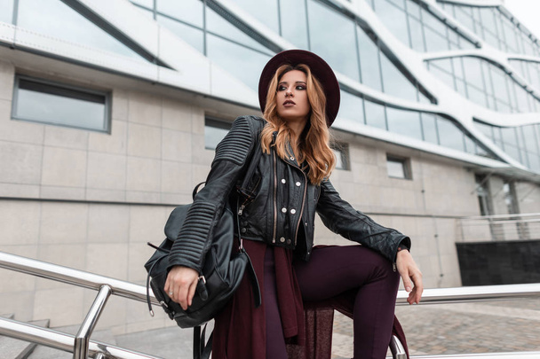 Moderni nuori punatukkainen nainen violetissa tyylikkäässä hatussa trendikkäässä nahkatakissa, jossa on musta tyylikäs reppu trendikkäissä housuissa lähellä kaupungin rakennusta keväällä. Seksikäs tyttö ulkona. Muoti
. - Valokuva, kuva