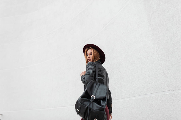 Jolie jeune femme élégante en violet chapeau élégant dans une veste en cuir vintage avec un sac à dos noir à la mode se promène près du mur blanc. Fille rousse moderne voyage. Mode printemps
. - Photo, image