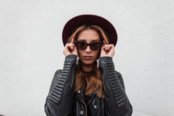 Retrato de mujer joven hipster de pelo rojo lujoso en gafas de sol oscuras de moda en sombrero púrpura en chaqueta de cuero elegante negro cerca de un edificio blanco al aire libre. Hermosa chica moderna modelo de moda
. - Foto, imagen