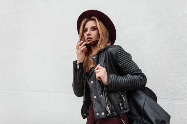 Modern pretty stylish hipster young woman in a fashionable hat in vintage dark sunglasses in a black jacket with a leather backpack in the city near white wall. European girl fashion model for a walk. - Photo, Image