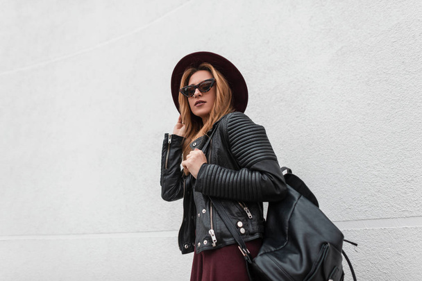 Sexy urban hipster young woman in a fashionable hat in stylish sunglasses in a vintage leather jacket with a backpack walks near white wall on a spring day. Modern red-haired girl travels. Youth style - Photo, Image