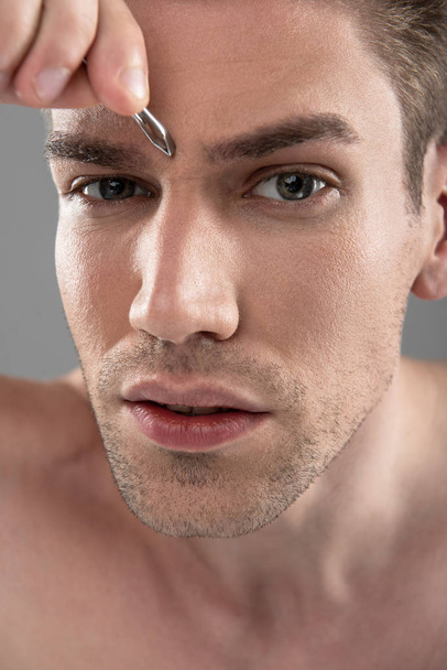 Attractive young man with stubble plucking eyebrows with tweezers - Foto, Bild
