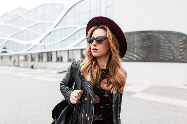 wunderschöne rothaarige Hipster junge Frau mit dunkler Sonnenbrille in einem eleganten Hut in einer stilvollen schwarzen Lederjacke mit einem trendigen Rucksack in der Stadt in der Nähe eines modernen Gebäudes. - Foto, Bild