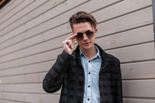 Elegante hombre hipster joven atractivo en gafas de sol oscuras de moda en una chaqueta a cuadros gris con un peinado de moda en una camisa cerca de una pared de madera en la ciudad. Guapo chico modelo disfruta de un paseo
. - Foto, Imagen