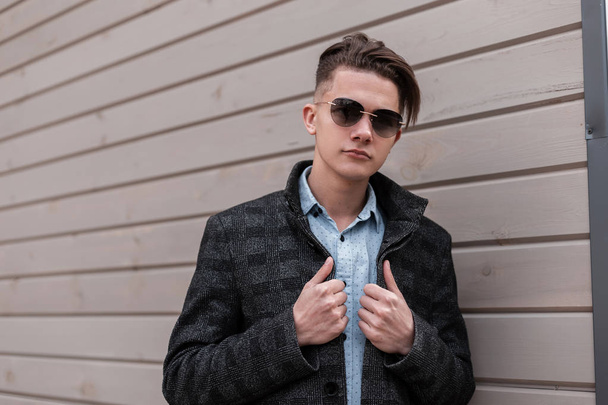 Mooie hipster jonge man in een stijlvolle geruite jas in een klassiek overhemd in modieuze zonnebrillen met een trendy kapsel poses in de buurt van een houten gebouw op de straat. Knappe knappe kerel. Herenkleding. - Foto, afbeelding