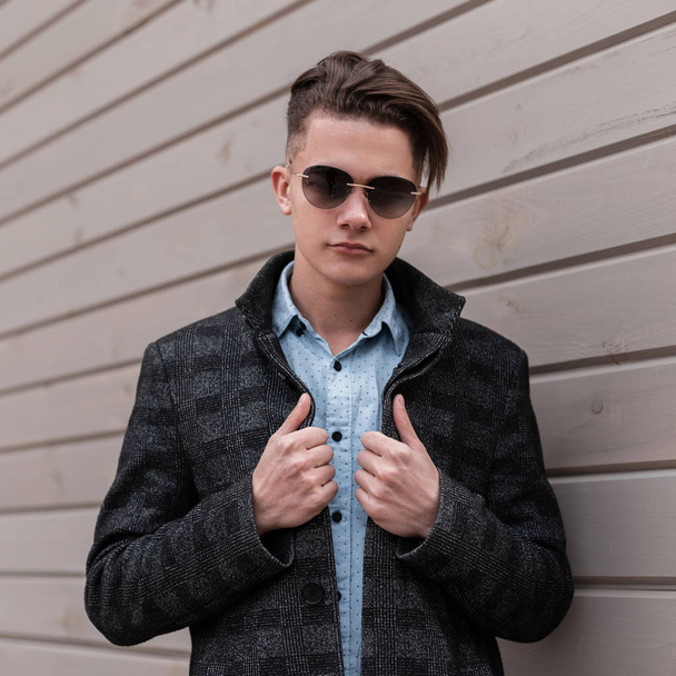 Serious handsome young hipster man in trendy sunglasses in a plaid jacket in an elegant shirt with a fashionable hairstyle stands near a vintage wooden wall in the city. Nice guy enjoying the weekend. - Fotó, kép