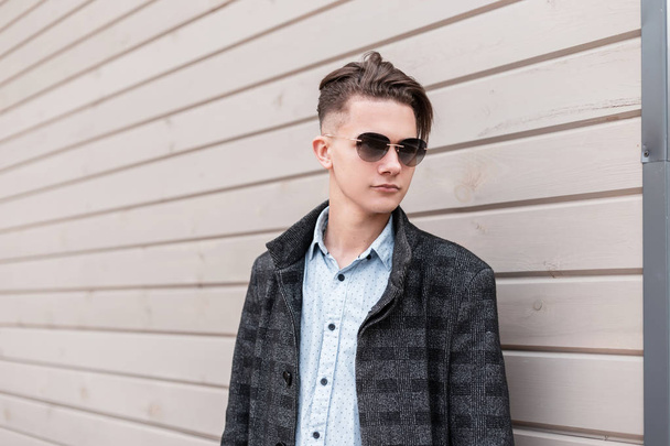 Knappe jonge hipster man met trendy kapsel in donkere zonnebril in een stijlvolle geruite jas in shirt poses op de straat in de buurt van een houten muur in de stad. Leuke kerel model buitenshuis. Moderne herenkleding. - Foto, afbeelding