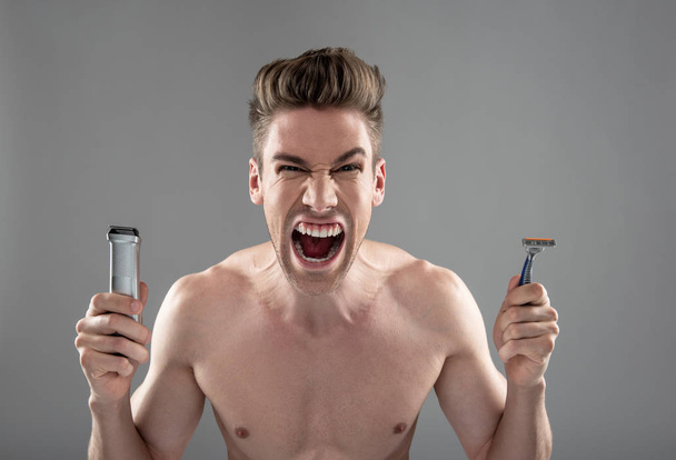 Angry young man holding electric shaver and disposable razor - Photo, image