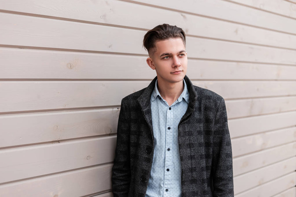 Handsome modern young man with a hairstyle in a gray checkered jacket in a stylish shirt in jeans is standing near a vintage wooden wall in the city on a spring day. Nice European guy outdoors. - Foto, immagini