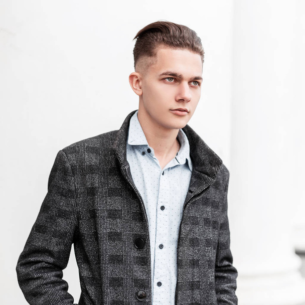 Handsome young man with a fashionable hairstyle in a vintage gray checkered jacket in a stylish shirt is standing near a white modern building in the city in the spring day. Attractive European guy. - Photo, Image
