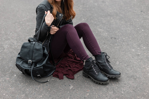 Young woman in a black stylish jacket in vintage purple pants in trendy leather boots with a leather backpack sits on the asphalt. New seasonal collection of fashionable women's shoes. Close-up. - Photo, Image