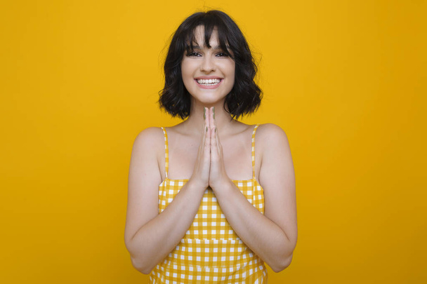 Portrait of a attractive brunette dressed in white dress show please with hands looking at camera smiling against a yellow studio wall. - Foto, Imagen