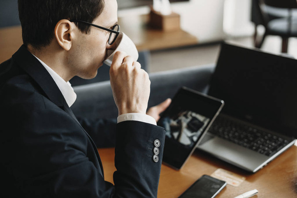Side view portrait of a young caucasian freelancer wearing glasses and dressed in suit drinking coffee and operating on a tablet in a coffee shop. - Foto, imagen