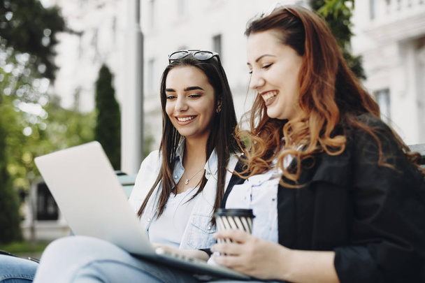 Όμορφη Καυκάσιος μελαχρινή με μακριά μαλλιά χαμογελά ενώ κοιτάζει το laptop φίλες της που είναι επίσης χαμογελά κάθεται σε ένα παγκάκι κατά ένα πολυτελές κτίριο. - Φωτογραφία, εικόνα