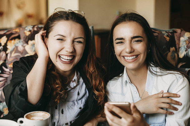 Portrét dvou úžasných přítelkyň, které se dívají na kameru, když popíjejí kávu v kavárně a používají telefony Smartphone. - Fotografie, Obrázek