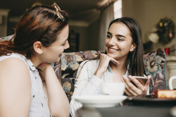 Mooie blanke vrouw met donker lang haar zittend in een coffeeshop glimlachend terwijl ze haar vriendin luisterde met een smartphone in haar handen. - Foto, afbeelding