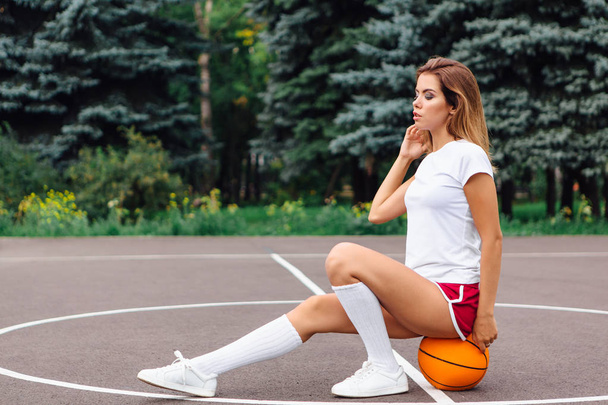 Beautiful young girl dressed in white t-shirt, shorts and sneakers, sits on a basketball court on the ball. - Fotó, kép