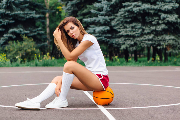 Menina bonita vestida com t-shirt branca, shorts e tênis, senta-se em um campo de basquete na bola
. - Foto, Imagem