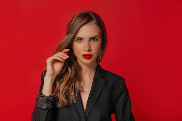 Модный портрет европейской привлекательной женщины с блестящими волосами и красными губами, смотрящей в камеру на красном фоне
  - Фото, изображение