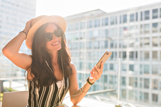 Bulanık kentsel arka planda çatıda akıllı telefon ile oturan hasır şapka koyu saçlı sevimli Avrupalı kadın - Fotoğraf, Görsel
