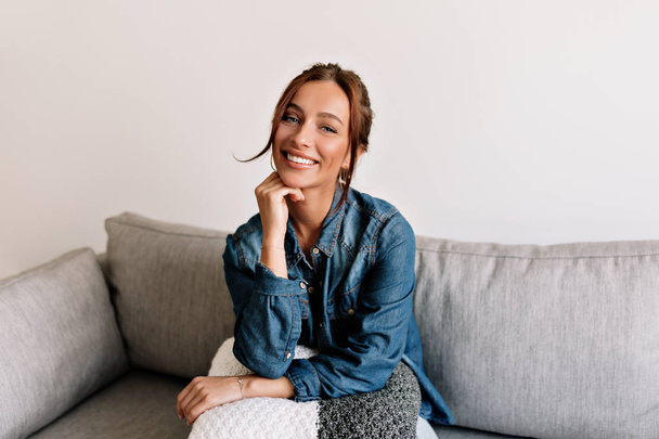 Menina rindo feliz com pele bronzeada e dentes brancos sentados no sofá no aconchegante apartamento e posando para a câmera
  - Foto, Imagem