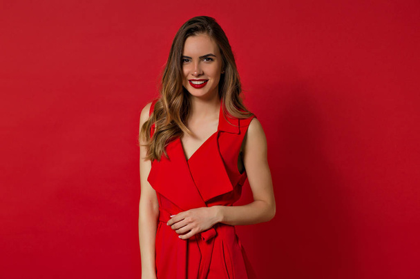 Smiling lovely woman with long hair and evening make up posing over red background and looking at camera - Photo, Image