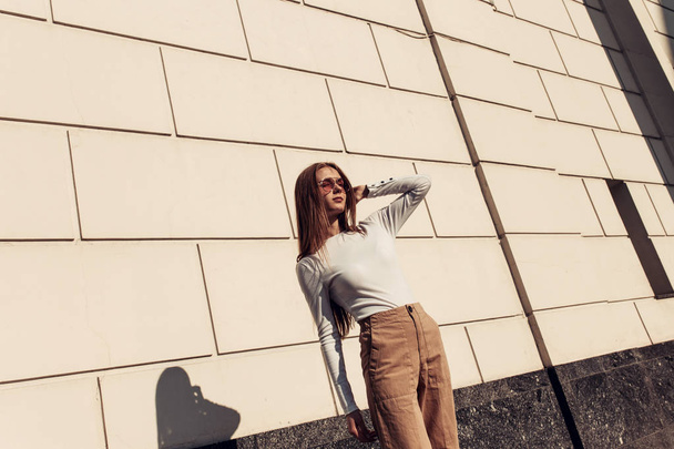 Fille posant contre un mur de briques blanches. Belle jeune femme portant un svidshot blanc et porte des lunettes de soleil
 - Photo, image
