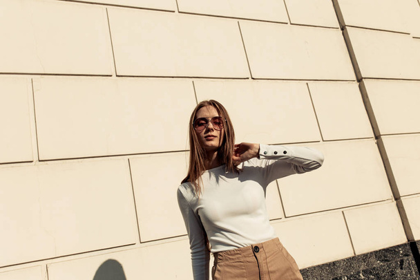 Beyaz tuğla duvara poz veren kız. Güzel genç kadın beyaz bir svidshot giyiyor ve güneş gözlüğü takıyor - Fotoğraf, Görsel