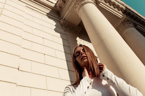 horizontal fashion portrait of a beautiful young woman with blond long hair against the background of high columns, sky and a brick wall. The girl looks down at the camera. Low angle shot. Sunlight - Foto, imagen