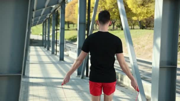 Portrait Of Fit Young Man With Jump Rope On Platform Near Metal Racks. Fitness Skipping Workout Outdoors. The Guy Jumps Near The Metal Pillars In The Background Of The Stadium. Dressed In A Black T - Кадри, відео