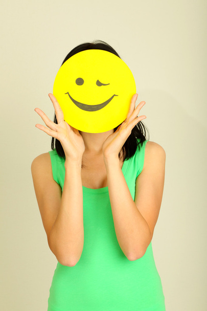 Young woman holding paper with happy smiley on gray background. - Foto, afbeelding