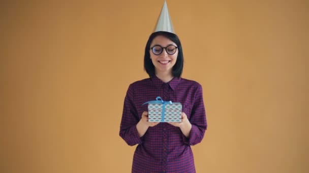 Portrait of cheerful young lady in bright party hat holding gift box and smiling - Felvétel, videó