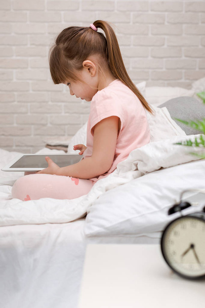 little child girl lies in bed uses digital tablet. - Фото, изображение