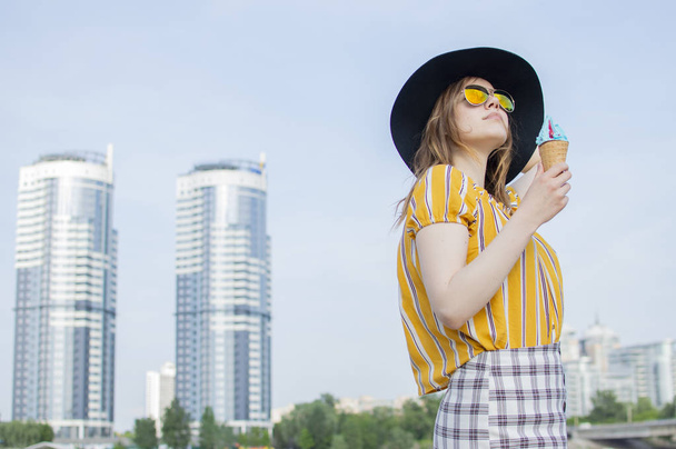 jovem menina bonita de pé na praia em uma camiseta listrada amarela em copos de laranja e um chapéu preto e comer sorvete, andando no verão fora da cidade
 - Foto, Imagem