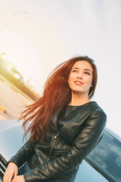 Gyönyörű bájos barna hosszú haja fiatal ázsiai nő, fekete bőr kabát közelében kocsiját - Fotó, kép