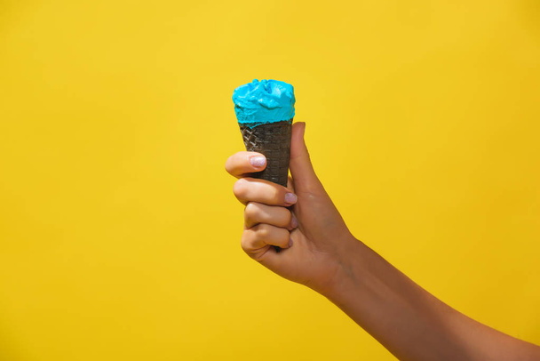 Sinistä jäätelöä vohvelikartiossa keltaisella pohjalla. Värillisen jäätelön sarvi naisen kädessä. Kylmä jälkiruoka värillisellä pohjalla
. - Valokuva, kuva