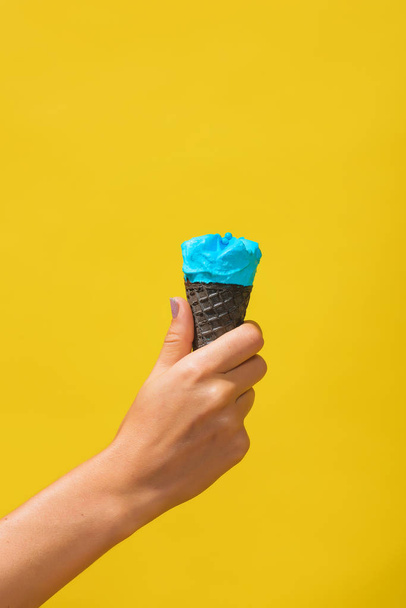 blaues Eis in einem Waffelkegel auf gelbem Hintergrund. Horn von buntem Eis in einer weiblichen Hand. kaltes Dessert auf farbigem Hintergrund. - Foto, Bild